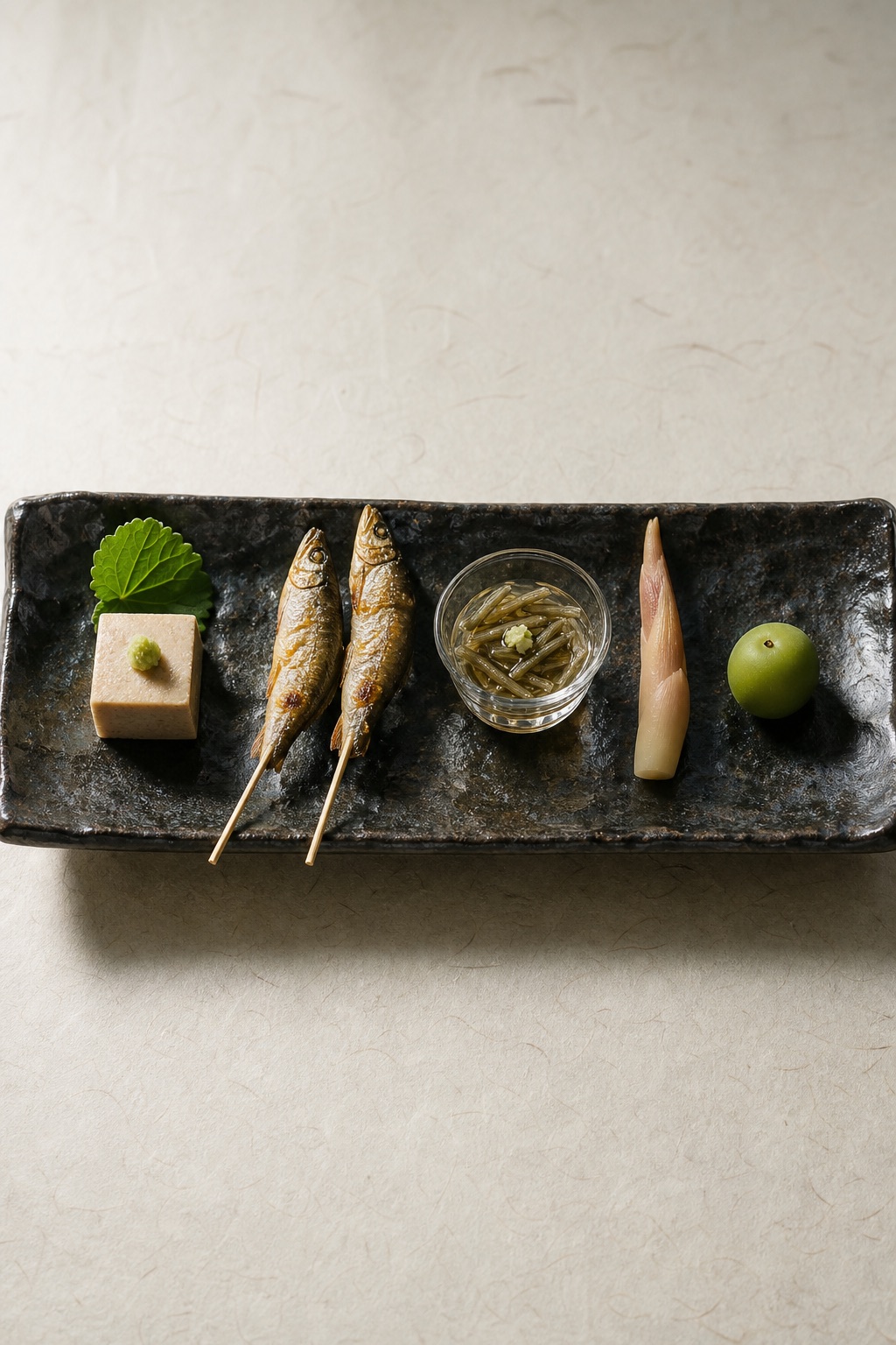 Overhead view of the hassun tray: five small summer bites arranged with wide space on dark glazed ceramic