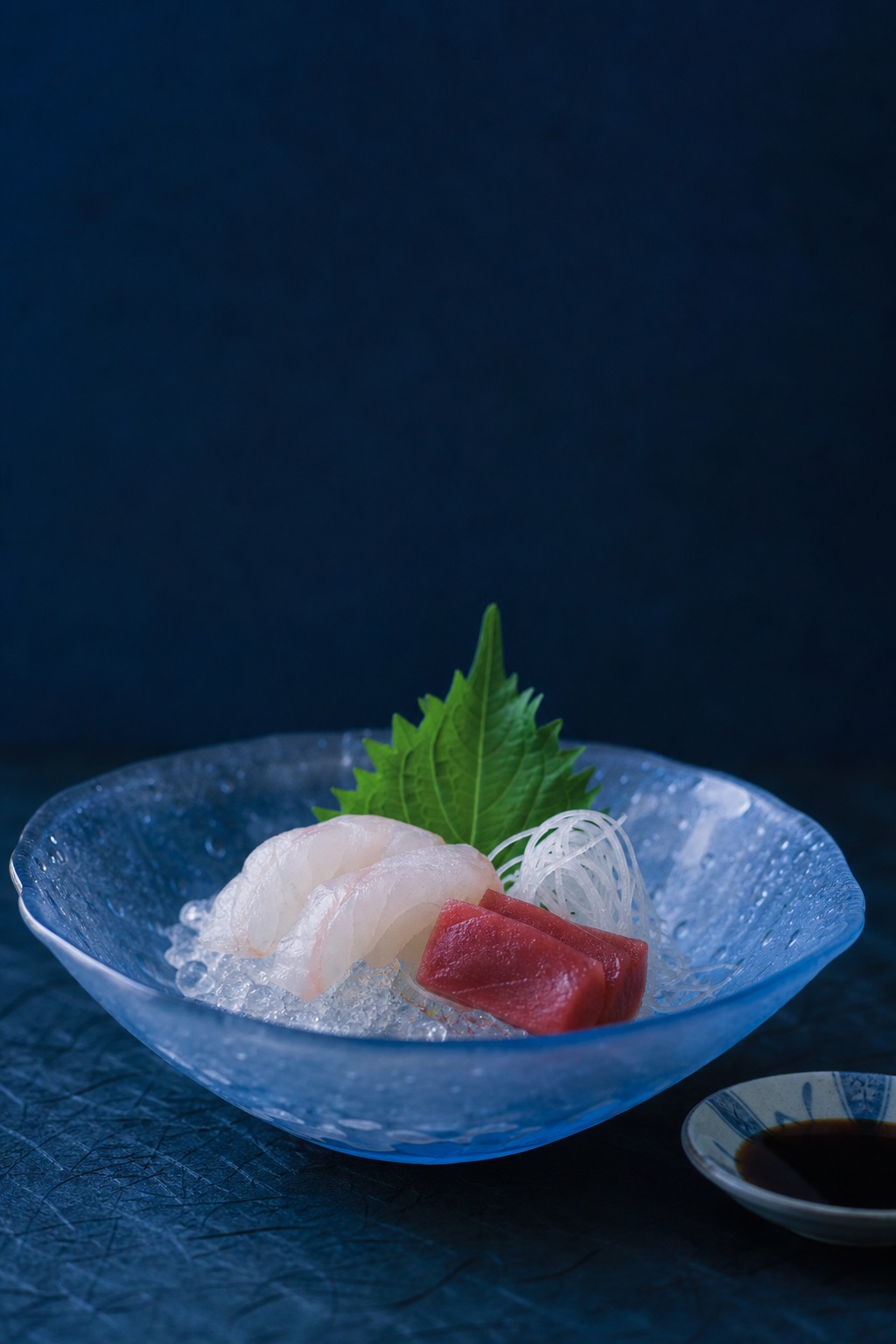 Sashimi of tilefish and tuna on shaved ice in a pale blue hand-blown glass bowl, condensation on the glass